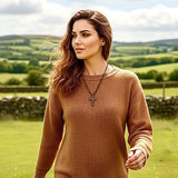 May include: A woman wearing a brown sweater and a cross pendant necklace. The sweater has a ribbed texture and long sleeves. The necklace features a dark metal cross. The background shows a green field and a cloudy sky.
