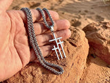 May include: Silver-toned necklace featuring a pendant with three stacked crosses. The chain is a braided style. The necklace is displayed against a backdrop of reddish-brown rocks and sand, with sunlight illuminating the scene.
