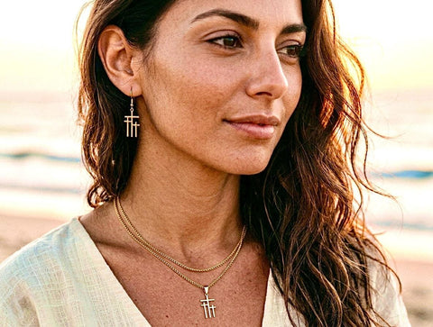 May include: A woman wearing gold-colored jewelry, including a necklace and earrings. The earrings and pendant feature a three-bar design. The necklace is a double-strand chain. The woman has long, brown hair and is wearing a cream-colored top.